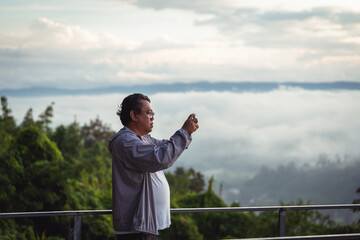 Man Standing on a Mountain Viewing Scenery while Using a Smartphone to Capture Pictures against a...