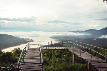 Obraz premium Serene Overlook of Misty Valley and River at Sunrise with Wooden Platform Surrounded by Lush Greenery and Mountains in Background