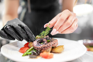 Chef plating gourmet octopus dish with roasted vegetables and fresh herbs. Close-up of hands adding thyme. Fine dining, culinary art, professional kitchen.