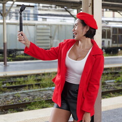 Asian woman stands on train platform in red coat and beret, smiling and holding an action camera on a selfie stick—stylish, confident, and framed by a casual urban transit setting