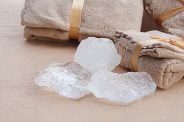 Natural alum crystal stones placed beside folded beige towels tied with ribbon on soft fabric background. The image represents natural spa, mineral purity, cleanliness, eco-friendly self-care products