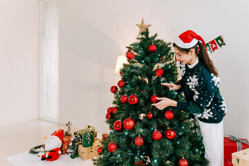 Woman in Santa hat decorates a Christmas tree with red ornaments in a bright modern living room, with gifts and festive decor creating a cozy holiday atmosphere.