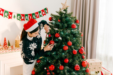 Woman in Santa hat decorates Christmas tree with red ornaments at home. Festive banner, sparkling...