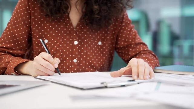 Close up of a female hand signing documents at a desk at a workplace in a modern business office. Businesswoman in a casual shirt checks the folder with documentation and signs the contract with a pen