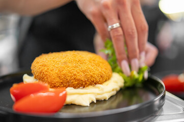 Close-up of a hand garnishing a plated dish with herbs. Fried patty on mashed potatoes, tomato wedges on the side. Professional food presentation in warm lighting