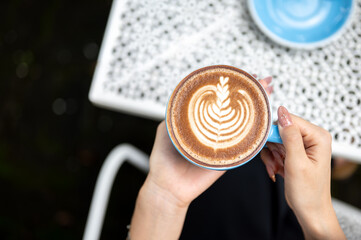 Close up a woman's hands holding latte art coffee in blue ceramic cup over floral pattern cafe table