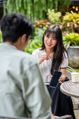 Pretty asian woman holding and showing phone to man while sitting on chair at table in cafe's garden