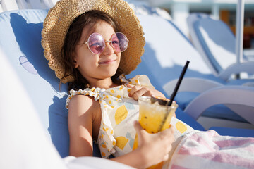 Funny cute girl drinks a cocktail on summer vacation. The child has fun on the beach. Cute baby in a colorful swimsuit and sunglasses is resting.