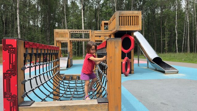 Little girl climbing rope net at modern wooden playground with slides on summer day in park surrounded by birch trees, concept of childhood and outdoor activity