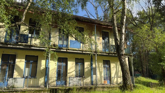Old abandoned two-story building with peeling paint and broken windows among trees, sunlight and shadows, concept of decay, history, forgotten places, concept of architecture and nostalgia
