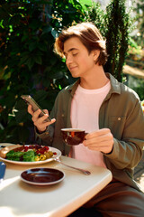 Fototapeta premium Young man enjoys a meal while checking his phone at a cozy modern cafe