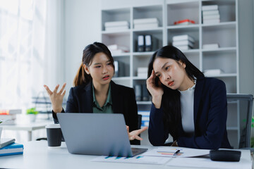 Two businesswomen argue during an office meeting, one visibly frustrated while the other clutches her head in stress, illustrating workplace communication breakdown and burnout