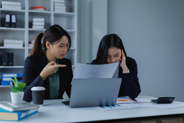 Two stressed business women are having a difficult conversation at their office desk, with one pointing at documents and the other appearing overwhelmed by pressure and a demanding boss