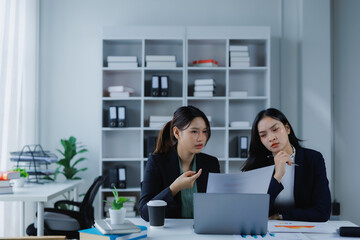 Two young businesswomen are collaborating, discussing a financial report and statistics, using a laptop during a meeting in a modern office environment
