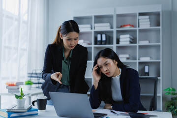 Businesswomen discussing a challenging project, one employee feeling stressed and holding her head while another points at the laptop screen, identifying an error in the workplace