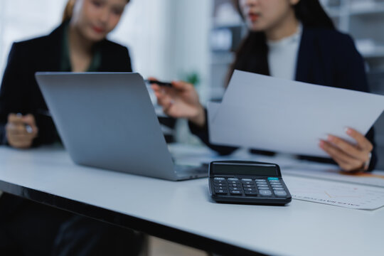 Two businesswomen are collaborating in an office, discussing financial documents and reports while using a laptop and calculator for budgeting, strategy, and planning data analysis