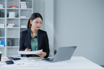 Asian businesswoman working diligently in a professional office environment, reviewing financial documents and utilizing a laptop for data analysis and accounting tasks