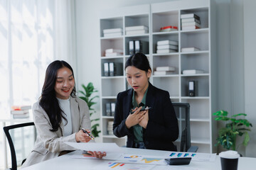 Two Asian businesswomen reviewing documents and financial charts together at an office desk, one showing a cheerful expression while the other reacts with a surprised yet relieved gesture