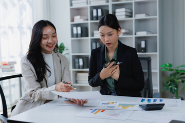Two young Asian businesswomen discussing financial data and charts in an office, one presenting surprising or exciting news to her colleague who reacts with an open mouth and hand to her chest