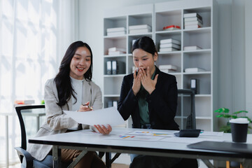 Two businesswomen discussing a report in an office, one sharing good news with a surprised and delighted colleague covering her mouth, indicating success and achievement