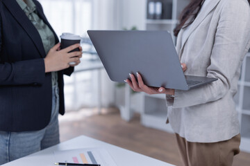 Businesswomen discussing work and analyzing data, using a laptop and holding a coffee cup, fostering collaboration and teamwork in a professional office environment