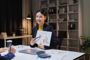 Asian businesswoman presenting sales charts and market growth to colleagues at a modern office desk, pointing to graphs while discussing data, strategy and team collaboration