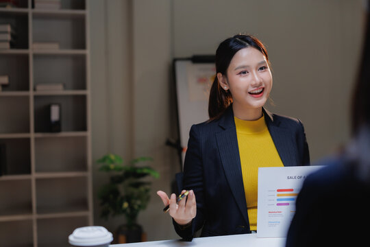 Young Asian businesswoman smiling and presenting a sales chart to a colleague during a professional corporate meeting, discussing business growth and strategy in the office