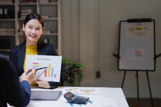 Asian businesswoman presenting financial data using a bar chart to a colleague, discussing product sales performance and analyzing market trends in a corporate office setting