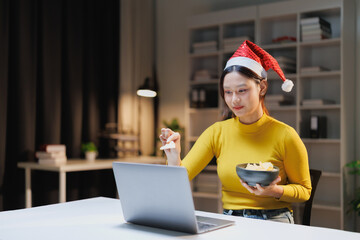 Young Asian woman wearing a Santa hat and yellow sweater enjoying snacks while participating in a virtual Christmas party or video call on a laptop at a home office desk at night