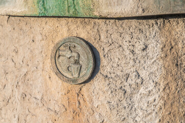 Runde Plakette aus Stein mit Abbildung eines Wasserhahns mit fließendem  Wasser in Trinkbecher an Brunnenwand mit grobem Verputz und grüner Patina an Trinkwasserbrunnen in Riederich