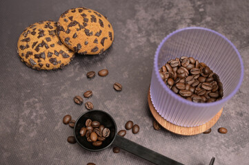 Dark roasted coffee beans in a transparent glass on a wooden board with a black spoon and oatmeal cookies with chocolate chips on the side. Dark background. Still life