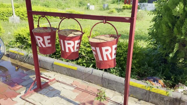 Three red fire buckets filled with sand hang from a metal stand in an outdoor area. A simple, traditional fire safety setup used in public places and industrial areas.