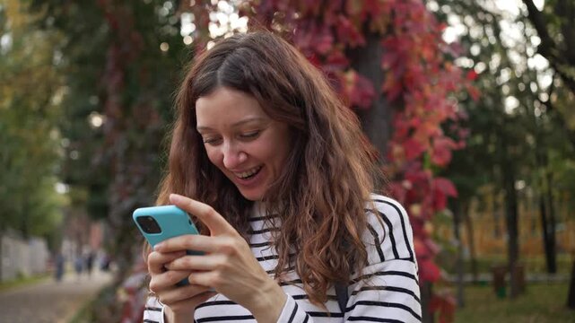 Cheerful young woman expressing shock and happiness while using her mobile phone in a beautiful autumn park, reacting to a surprising message or unexpected notification on the screen