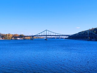 Obraz premium Sunny autumn day on the Dnipro River embankment in Kyiv. View of the river, bridges, and modern urban development on the left bank. Bright blue sky and water.
