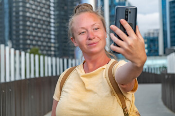 Young woman taking a selfie while enjoying a sunny day in a modern city setting with tall buildings...