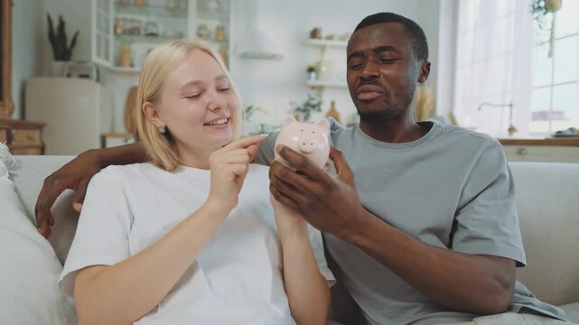 Cheerful interracial loving couple holding piggy bank and coins in hands. Beautiful blonde woman and her boyfriend discussing future purchases, save money for future, financial literacy and savings - Powered by Adobe