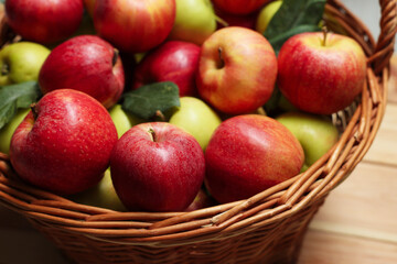 Fresh apples in wicker basket and green leaves on wooden table, closeup