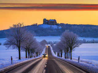 Obraz premium A captivating winter landscape features a car driving on a long, snow-covered road flanked by frosty trees, leading towards a majestic castle on a distant hill under a vibrant orange and yellow sunris