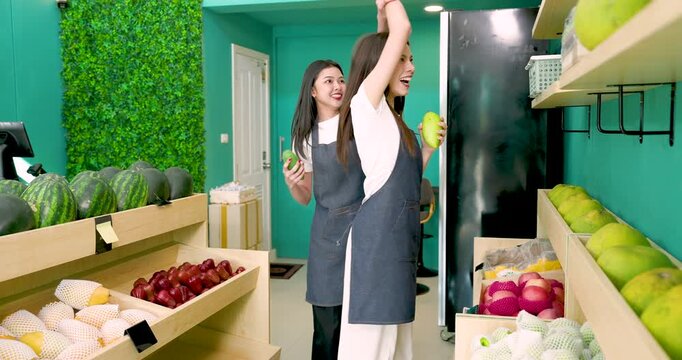 Asian woman and caucasian woman wearing aprons raise hands and spin joyfully while holding fruit, dancing together happily inside local fruit store before welcoming customers to small business