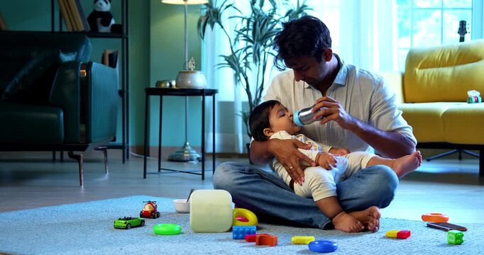 Indian infant baby boy drinking milk from bottle and father feeding while sitting on carpet in bright modern home, showcasing loving parenting, nurturing bond, care, and joyful family moments indoors