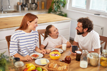 A woman and a child share a joyful breakfast at the kitchen table with a man, surrounded by various foods and warm atmosphere before starting the day.