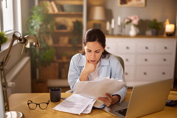 A woman is sitting at a wooden desk in her home office, reviewing financial papers while appearing concerned about her current financial situation.