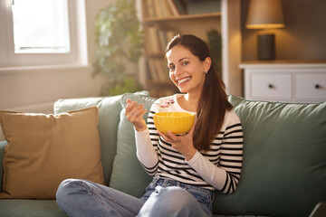 A woman smiles while eating cereal from a bright yellow bowl in a relaxed living room setting. Natural light filters through the window, creating a warm atmosphere. © Stockphotodirectors