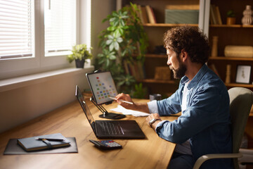 Individual engages in productive activities at a well-organized home office, utilizing a laptop and tablet, surrounded by greenery and soft natural light.