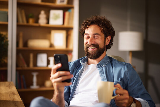 A man with curly hair and a beard sits in a comfortable home office, smiling while holding a smartphone in one hand and a cup of coffee in the other. Sunlight illuminates the space.