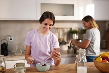 A young girl enjoys her breakfast while looking at her smartphone. Her mother is preparing tea in the background, creating a warm family atmosphere.