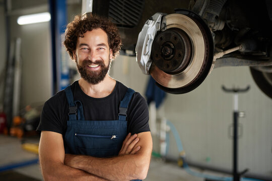 A skilled car mechanic with a beard stands in a well-equipped workshop. He is smiling while surrounded by tools and vehicle parts, ready to assist customers with car repairs and maintenance. - Powered by Adobe