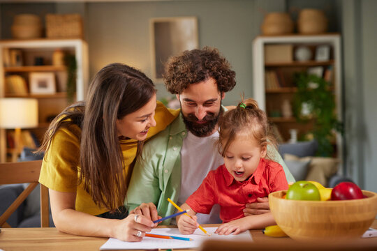 A family spends quality time at home, engaging in fun coloring and drawing activities at a table. Parents assist their child who is excitedly creating art. - Powered by Adobe