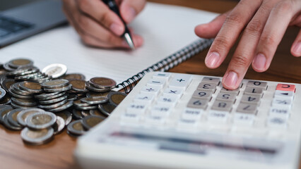 Expense tracking and saving money with calculator and coins, Close-up hand pressing calculator with pile of coins and notebook, saving money, and monthly budget management for finance planning.