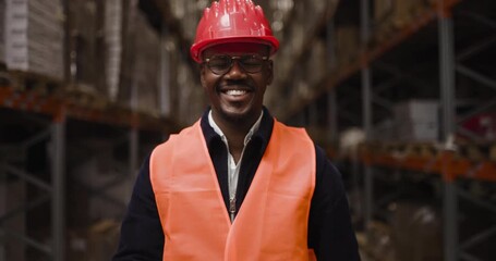 Confident warehouse worker smiling towards the camera, standing in an industrial storage aisle, wearing safety gear including a hard hat and reflective vest, representing logistics and supply chain - Powered by Adobe